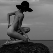 Wife, face hidden by hat, kneels nude on a large stone.
